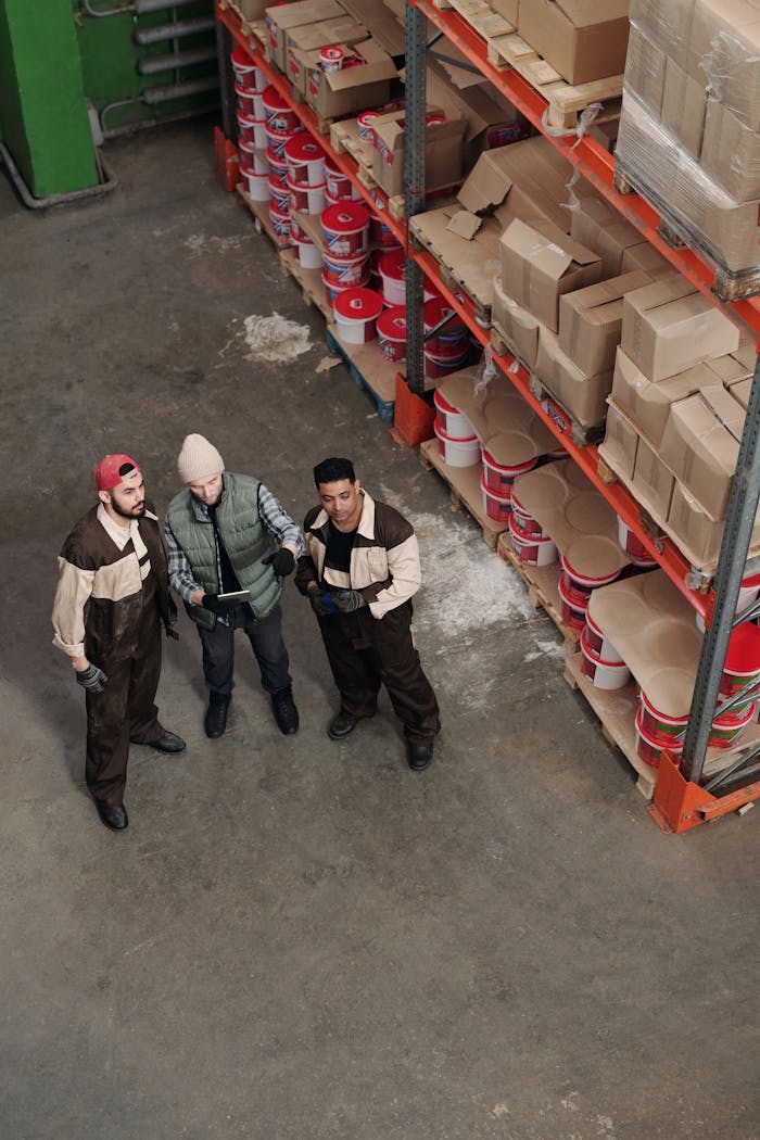 Three workers in a warehouse discussing inventory management near shelves with boxes and paint cans.