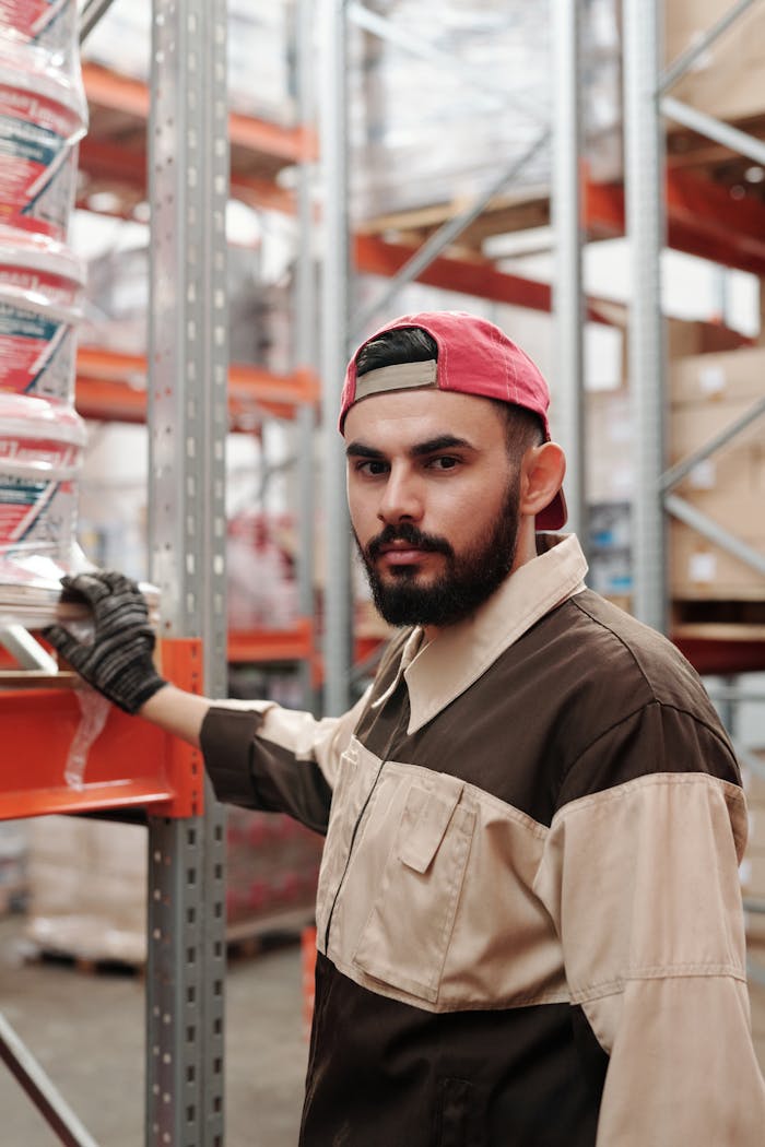 Portrait of a bearded man in a warehouse wearing a red cap and work uniform.